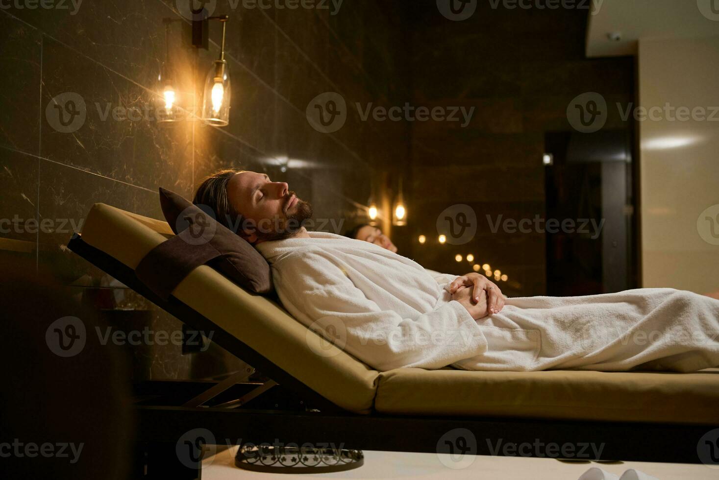 atraente bonito meio envelhecido homem dentro roupão de banho relaxante com fechadas olhos, deitado em uma chaise salão perto uma borrado mulher em a fundo, dentro uma luxo sereno atmosfera do uma bem estar spa Centro foto