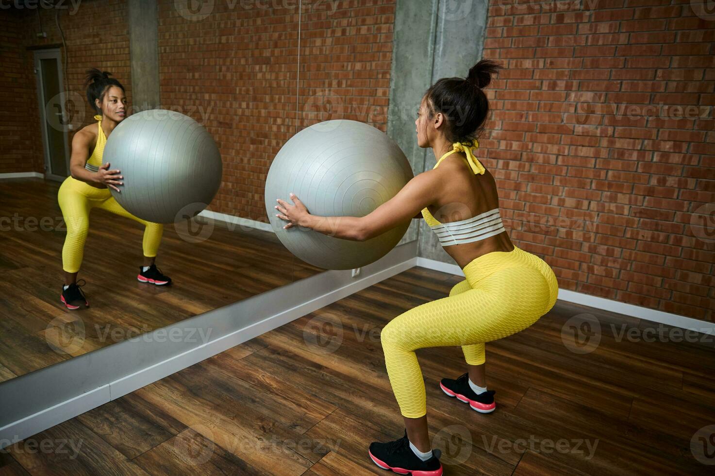 traseiro Visão do uma desportivo mulher segurando uma cinzento ginástica bola e fazendo glúteo exercícios, bombeamento nádegas enquanto agachado dentro frente do uma espelho dentro ginástica estúdio foto