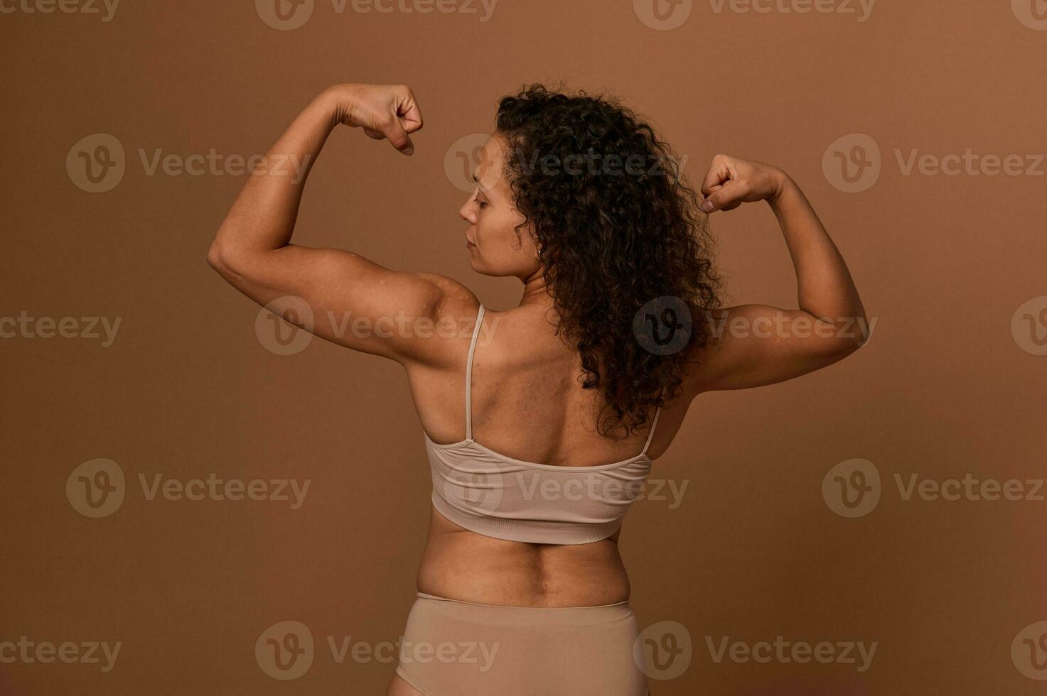 traseiro Visão do uma Deleitado confiante meio envelhecido africano americano mulher dentro bege roupa íntima olhando às dela braços e cerrado punhos, posando contra colori fundo com cópia de espaço. foto