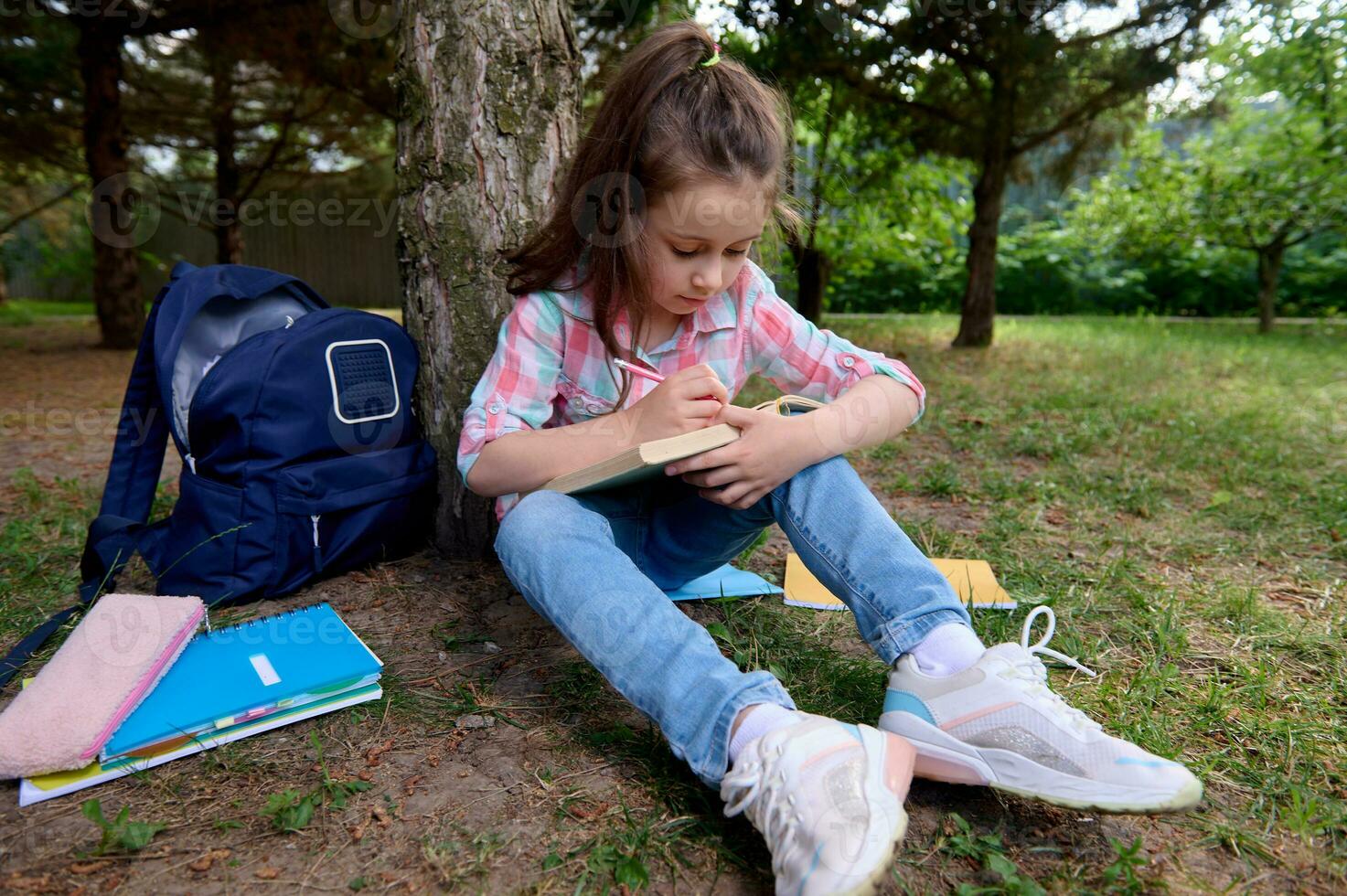 caucasiano adorável pequeno criança garota, uma primeiro motoniveladora escrevendo em pasta de trabalho, acionado em fazendo dever de casa depois de classe ao ar livre. foto