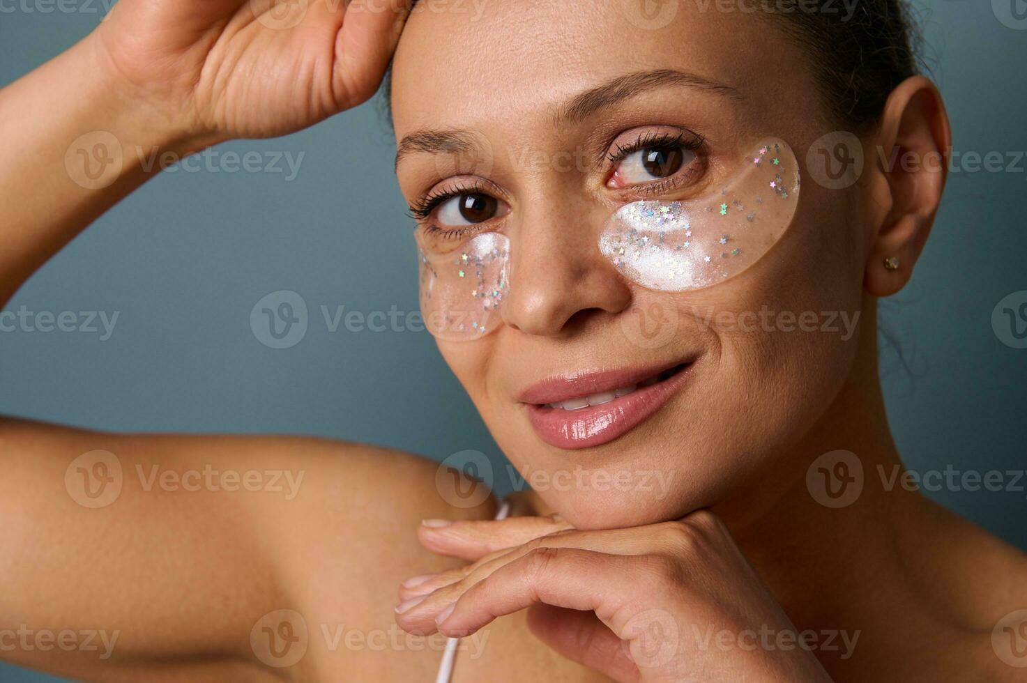 fechar-se retrato do a atraente sorridente mulher aplicando ouro médico hidrogel colágeno olho remendos para reduzir bolsas debaixo dela olhos e inchaço, posando às Câmera contra uma cinzento fundo. cópia de espaço foto