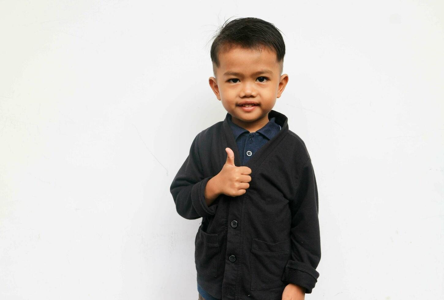 uma sorridente criança pequena Garoto parece às a Câmera levantando dele mãos para mostrar polegares acima, isolado em branco fundo foto