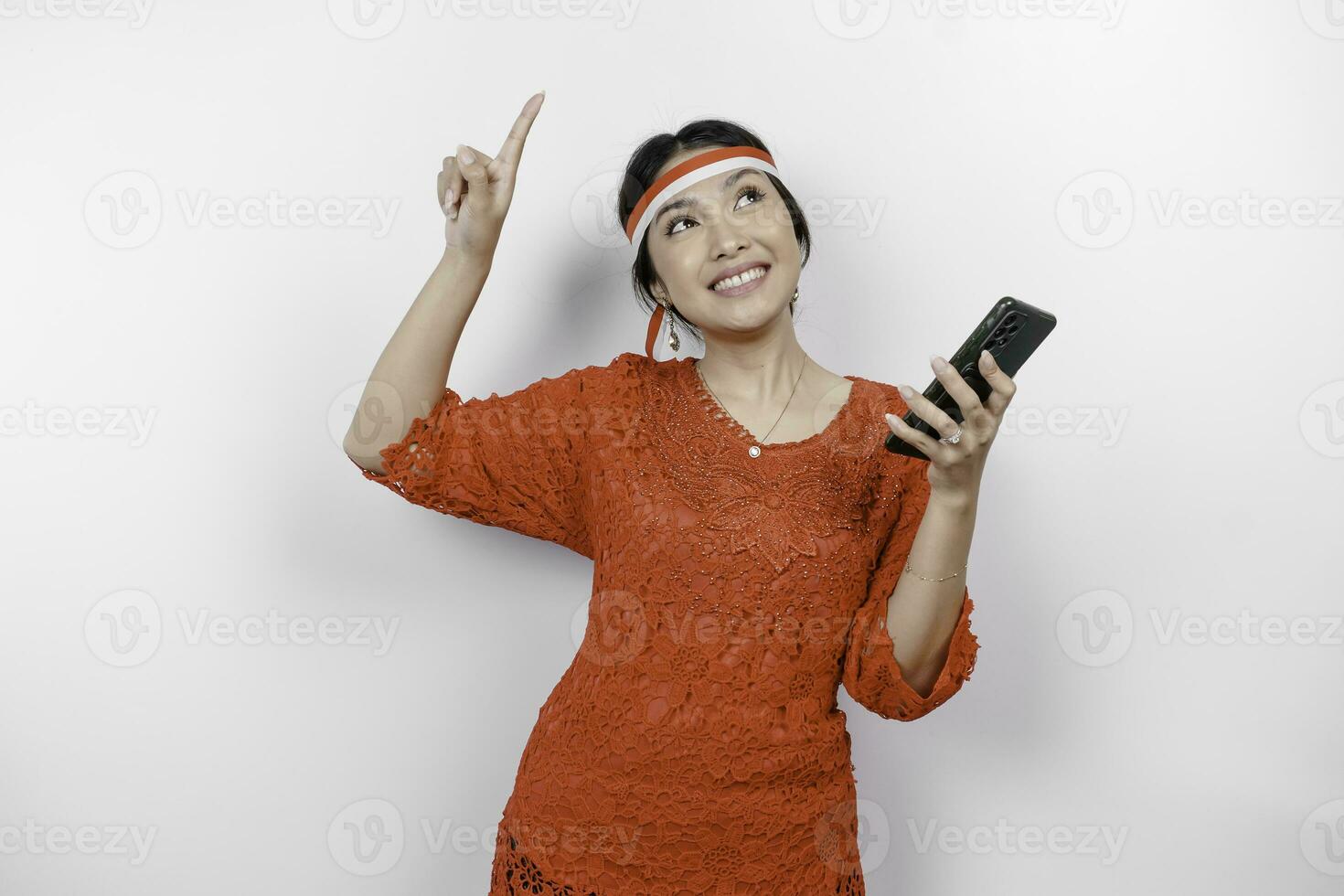 uma feliz ásia mulher vestindo vermelho kebaya e arco de cabelo, segurando dela telefone, e apontando cópia de espaço em topo do dela, isolado de branco fundo. da indonésia independência dia foto