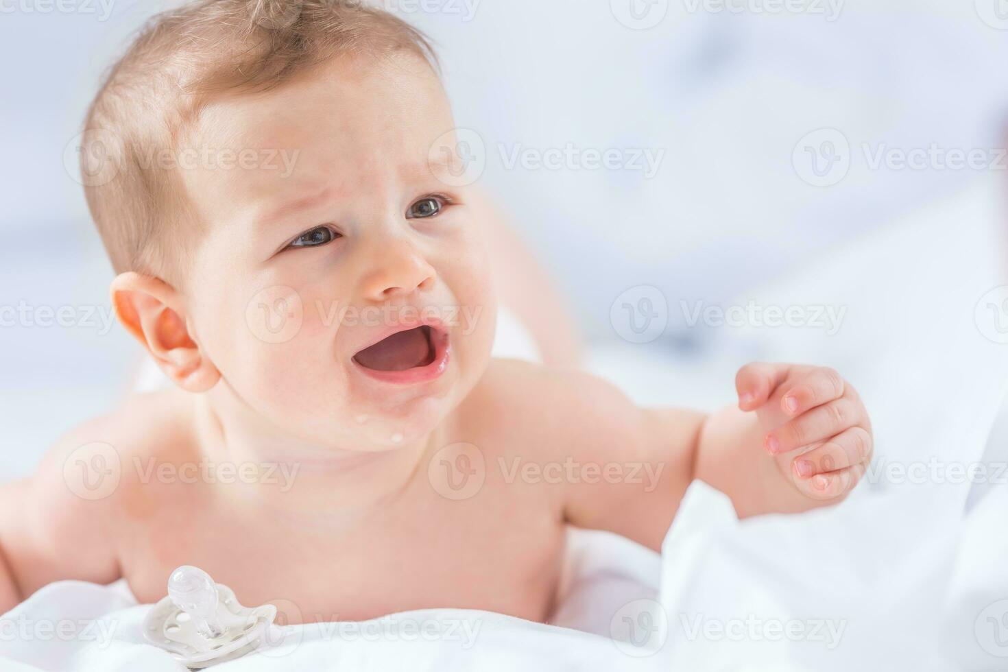 retrato do fofa criança pequena Garoto deitado em cama. recém-nascido criança dentro quarto. bebê mentiras em dele barriga levanta a cabeça e chorando foto