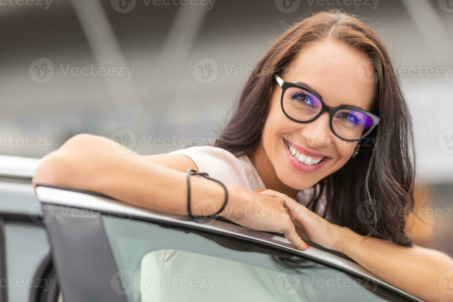 feliz Novo fêmea carro proprietário inclina-se em a aberto porta do a veículo com uma grande sorrir foto