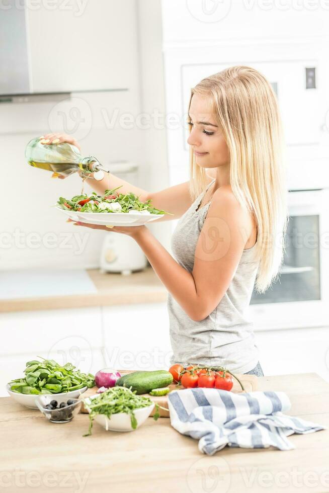 jovem feliz Loiras menina comendo saudável salada a partir de Rúcula espinafre tomates azeitonas cebola e derramando Oliva óleo foto