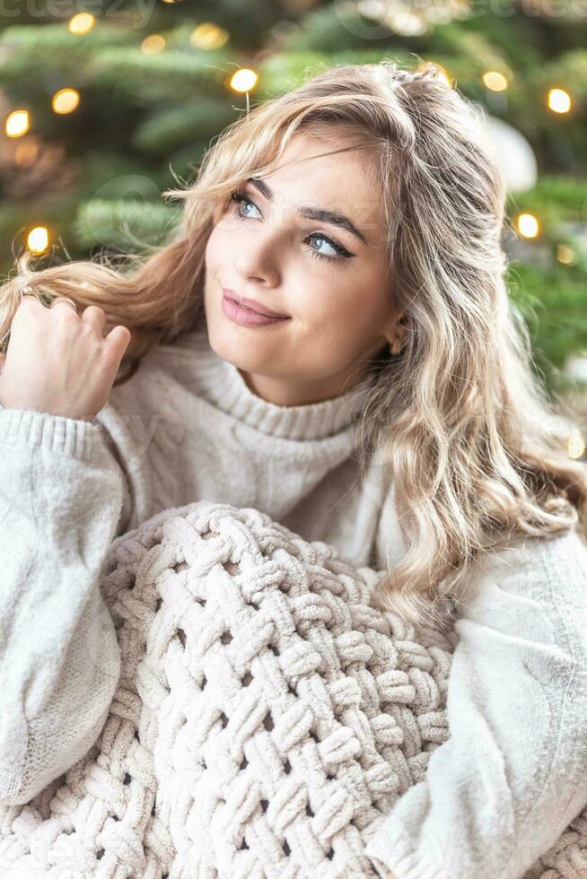 retrato do uma encaracolado Loiras menina dentro branco suéter dentro frente do uma Natal árvore foto