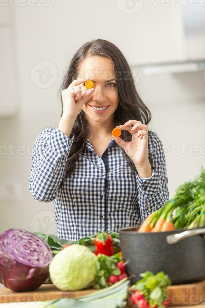 brincalhão jovem mulher segurando fatias do cenoura dentro a cozinha - dieta vegetal e heath conceito foto