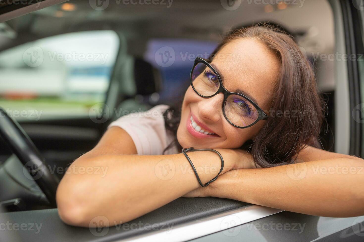 sorridente fêmea motorista vestindo óculos inclina-se através a aberto porta janela sentado em motorista assento foto