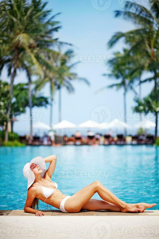uma mulher dentro uma branco chapéu e bikini deitado em a Beira do uma piscina foto