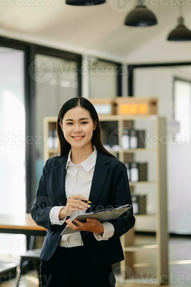 confiante o negócio especialista atraente sorridente jovem mulher segurando digital tábua em escrivaninha dentro escritório. foto