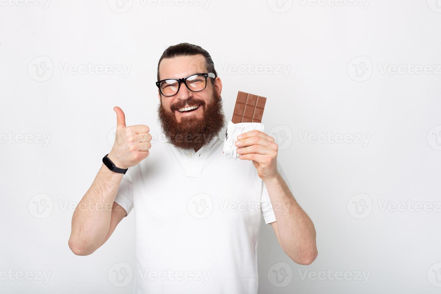 retrato de um homem barbudo alegre em uma camiseta branca mostrando o polegar para cima e uma barra de chocolate foto