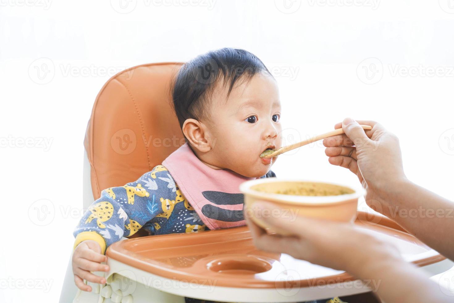 Menina de 6 meses de idade comendo mistura de comida em uma cadeira alta. foto