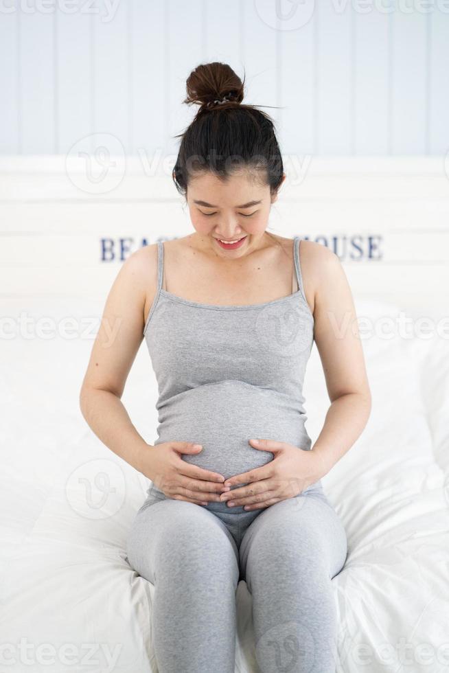 mulher grávida sentada na cama e tocando a barriga em casa. foto
