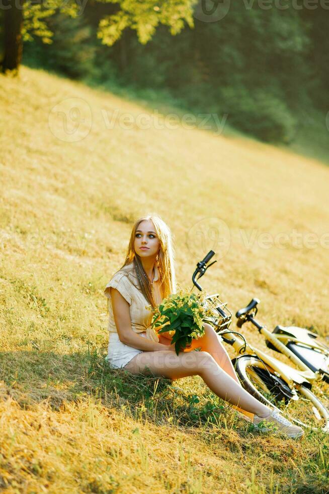 retrato do uma lindo menina dentro a floresta, sentado em a grama, Próximo para uma bicicleta, com uma ramalhete do flores, atrás a raios do a sol, dentro uma amarelo camisa e branco shorts, uma verão andar. foto