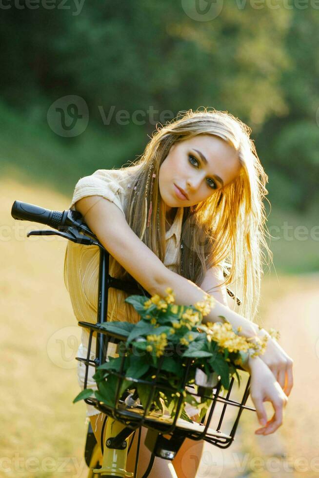 uma jovem lindo mulher detém uma bicicleta com amarelo verbena flores dentro uma cesta em a ao ar livre país andar foto
