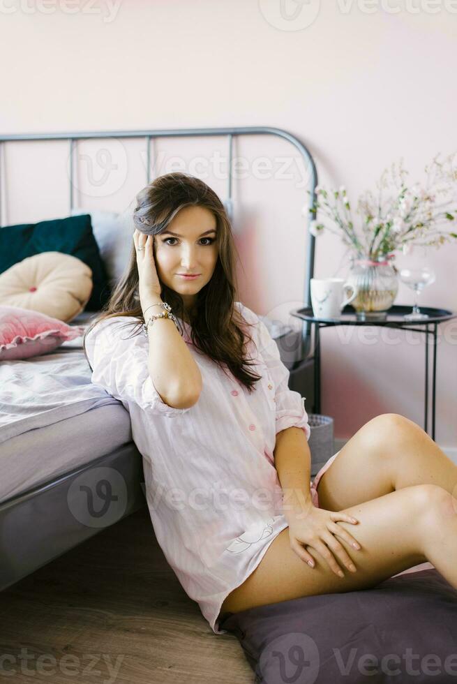 fofa caucasiano jovem mulher dentro pijamas é sentado em a chão perto a cama dentro a quarto e desfrutando dela vida foto