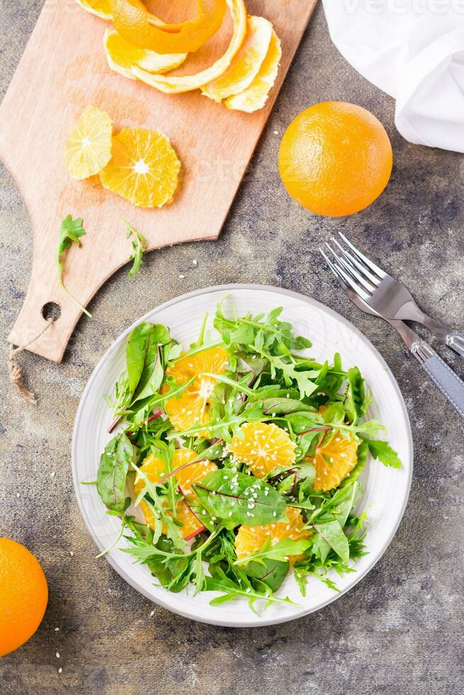 dieta vegetariano Vitamina salada do laranja fatias e uma misturar do Rúcula, acelga e mizun folhas em uma prato e uma corte borda com descascado laranja em a mesa. topo e vertical Visão foto