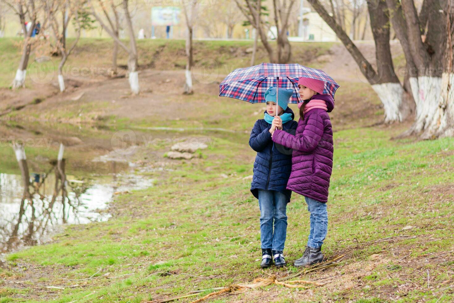 dois meninas irmãs ficar de pé debaixo a guarda-chuva em a costa do uma lago dentro cedo Primavera. andar ao ar livre foto