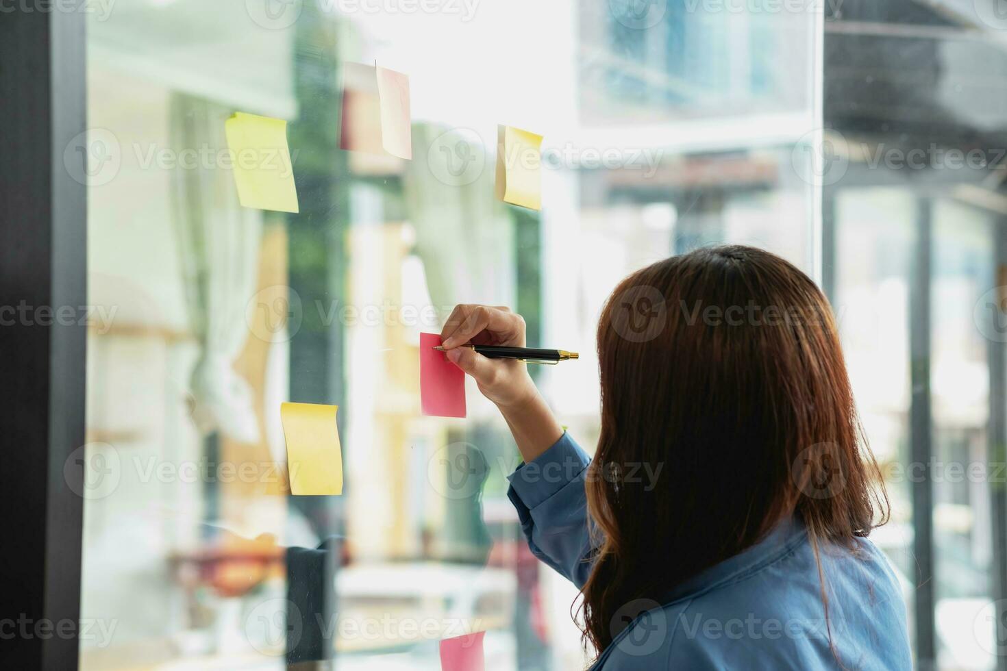 uma fêmea empregado escreve em uma colorida bloco de anotações. étnico mulheres trabalhando às iniciantes chuva de ideias colaborativo planos em vidro parede adesivos Vejo menos. foto