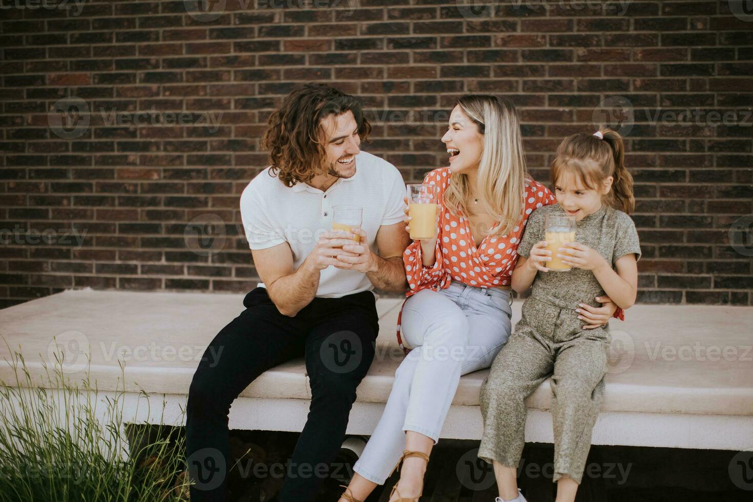 família com uma mãe, pai e filha sentado lado de fora em a passos do uma frente varanda do uma tijolo casa foto