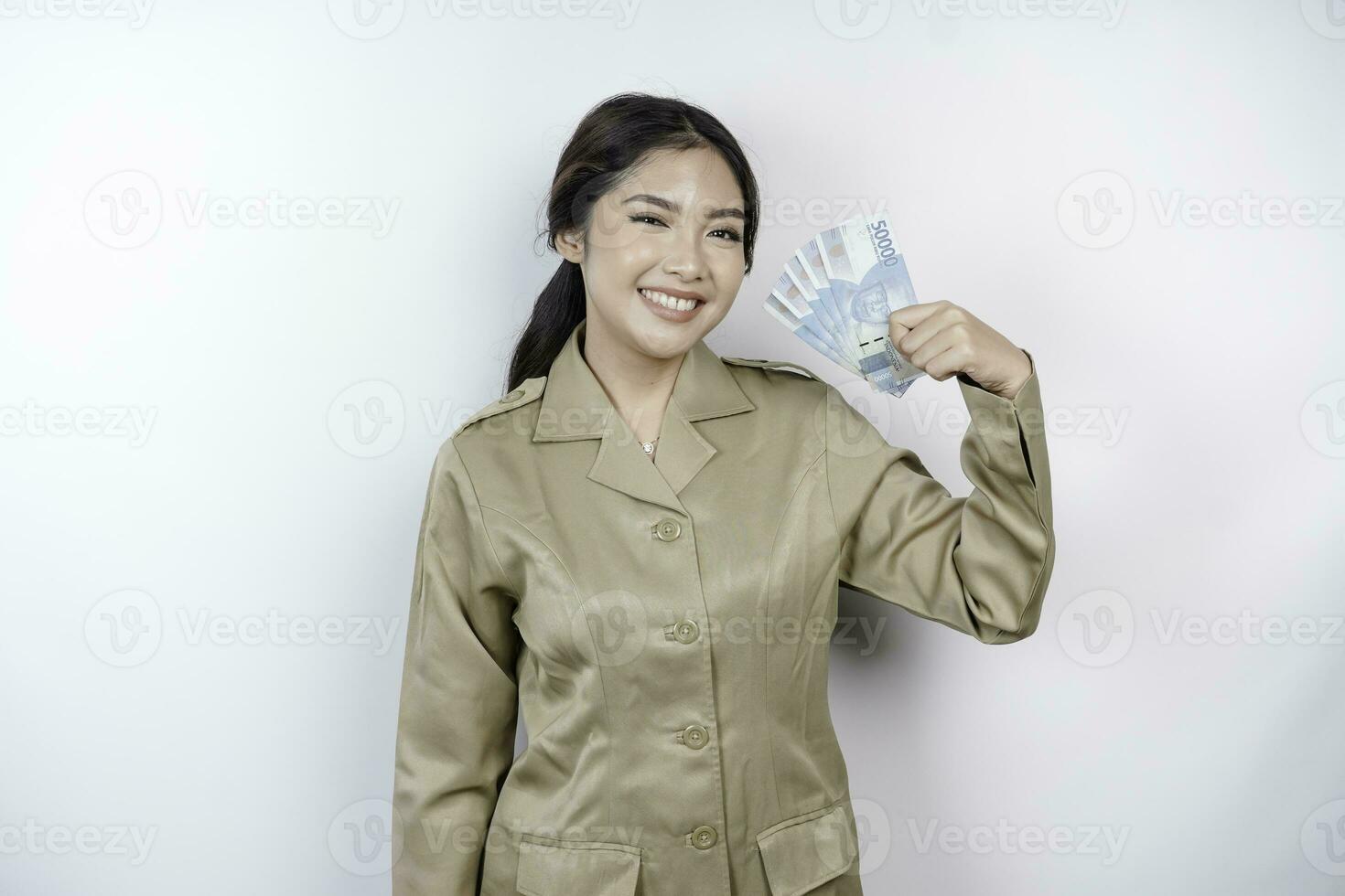 uma sorridente jovem governo trabalhador mulher é vestindo cáqui uniforme segurando dinheiro dinheiro dentro indonésio rupia isolado de branco fundo foto