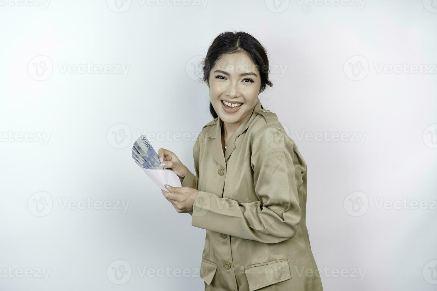 uma sorridente jovem governo trabalhador mulher é vestindo cáqui uniforme segurando dinheiro dinheiro dentro indonésio rupia isolado de branco fundo foto