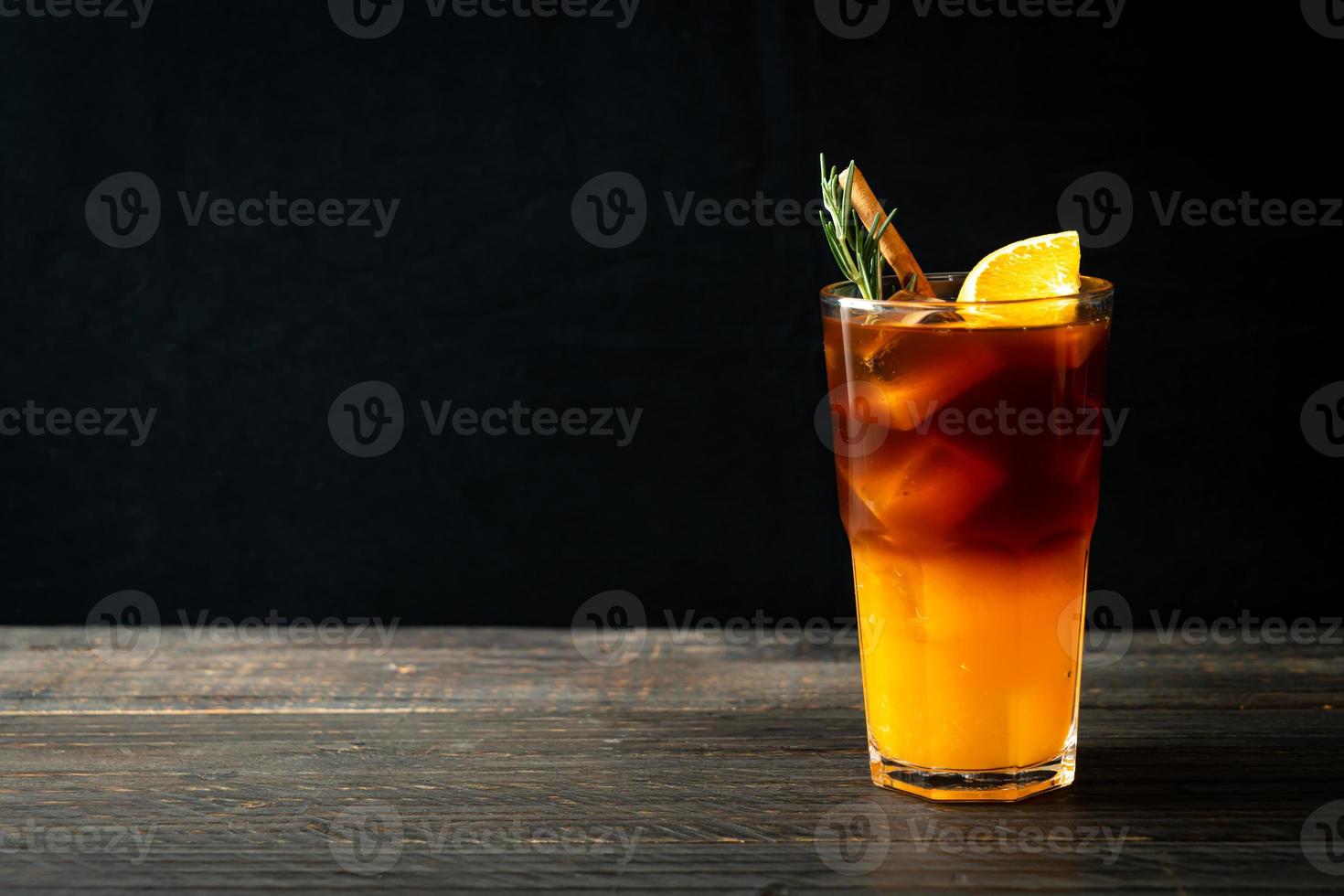 um copo de café preto americano gelado e uma camada de suco de laranja e limão decorado com alecrim e canela em um fundo de madeira foto