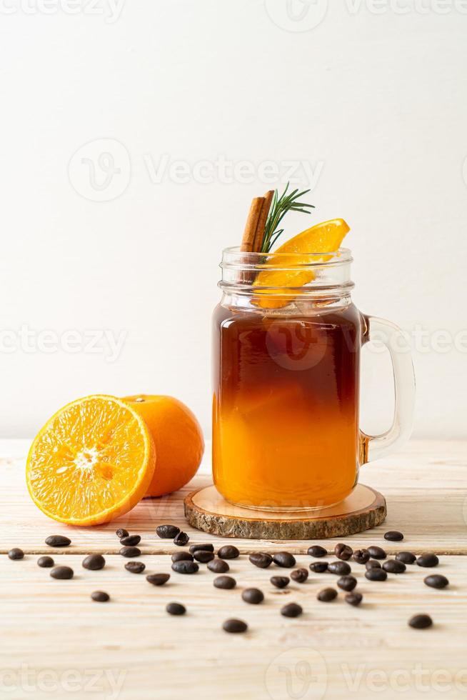 um copo de café preto americano gelado e uma camada de suco de laranja e limão decorado com alecrim e canela em um fundo de madeira foto