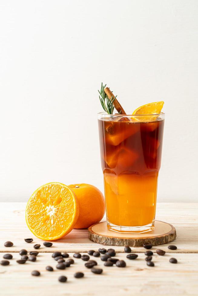 um copo de café preto americano gelado e uma camada de suco de laranja e limão decorado com alecrim e canela em um fundo de madeira foto