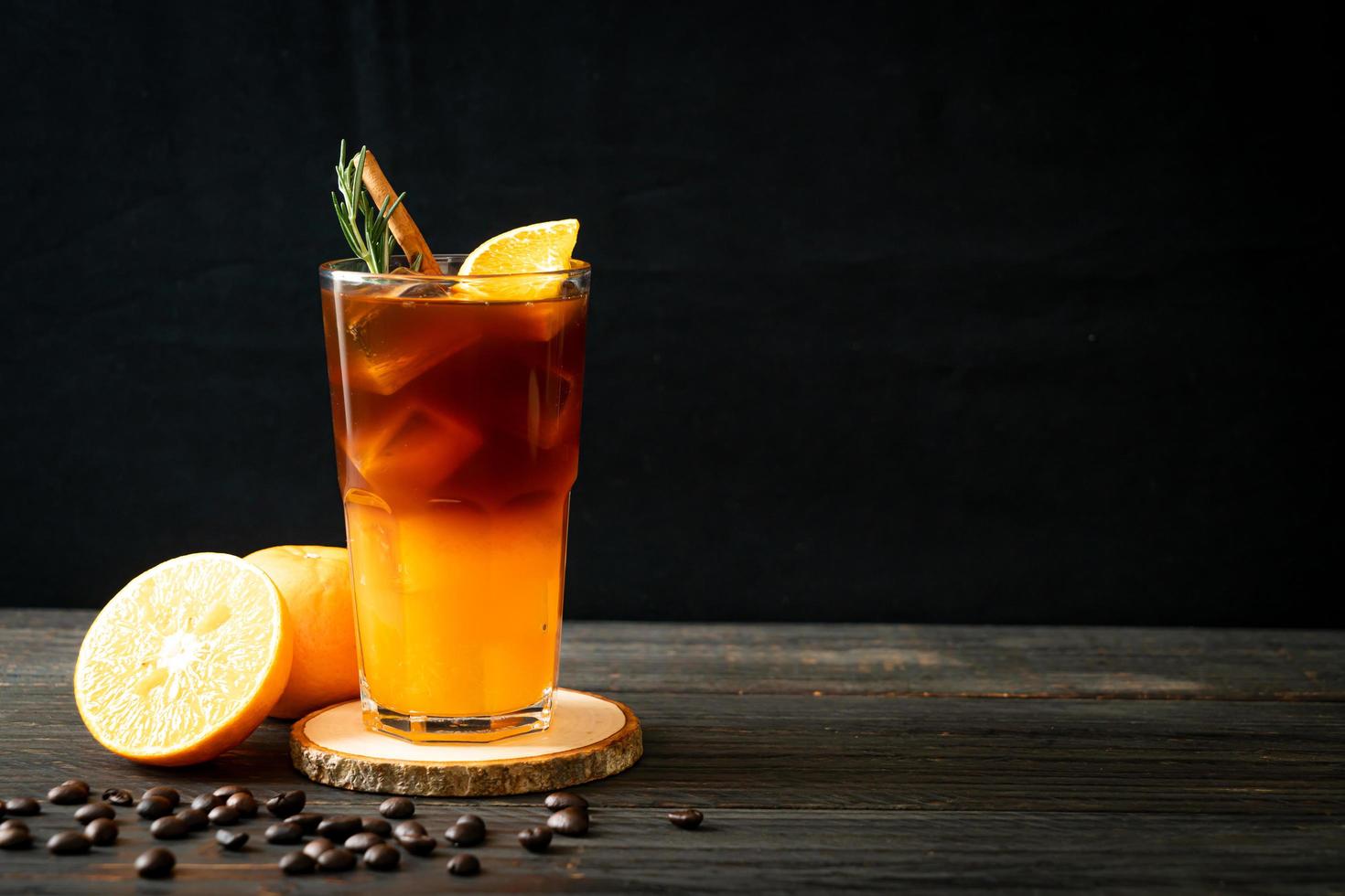 um copo de café preto americano gelado e uma camada de suco de laranja e limão decorado com alecrim e canela em um fundo de madeira foto