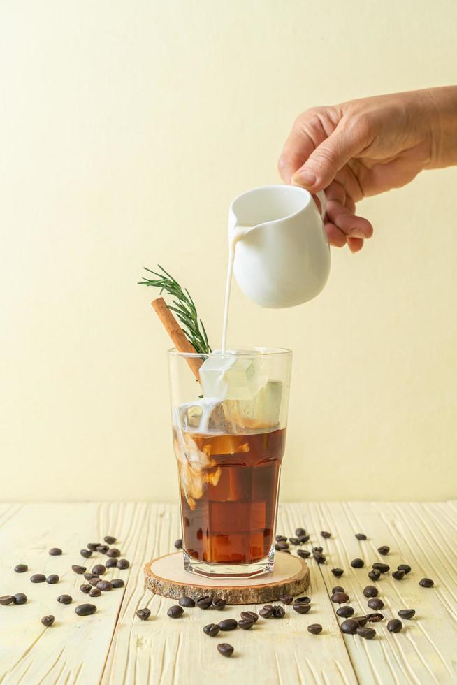 derramando leite em um copo de café preto com cubos de gelo, canela e alecrim no fundo de madeira foto