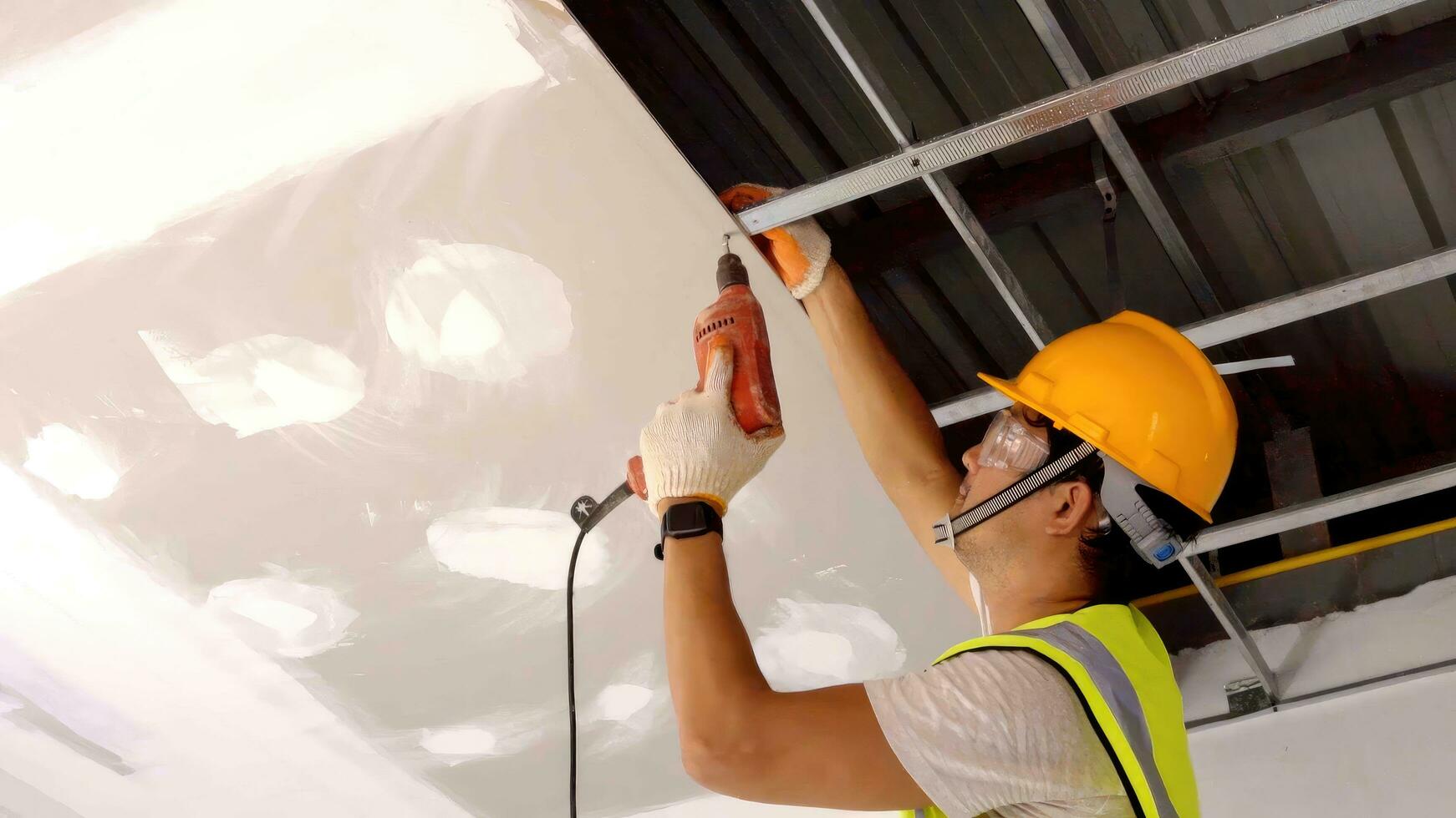 ásia construção trabalhador dentro segurança roupas e trabalhos luvas é fixação a drywall teto para a metal quadro, Armação usando a elétrico Chave de fenda em a teto cobrir foto