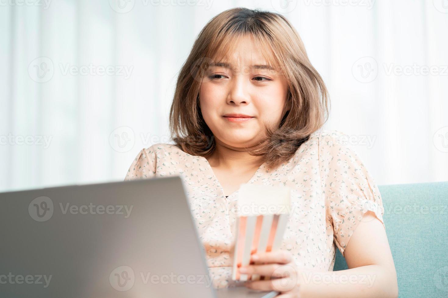 jovem asiática sentada no sofá, comendo e assistindo filmes no laptop foto