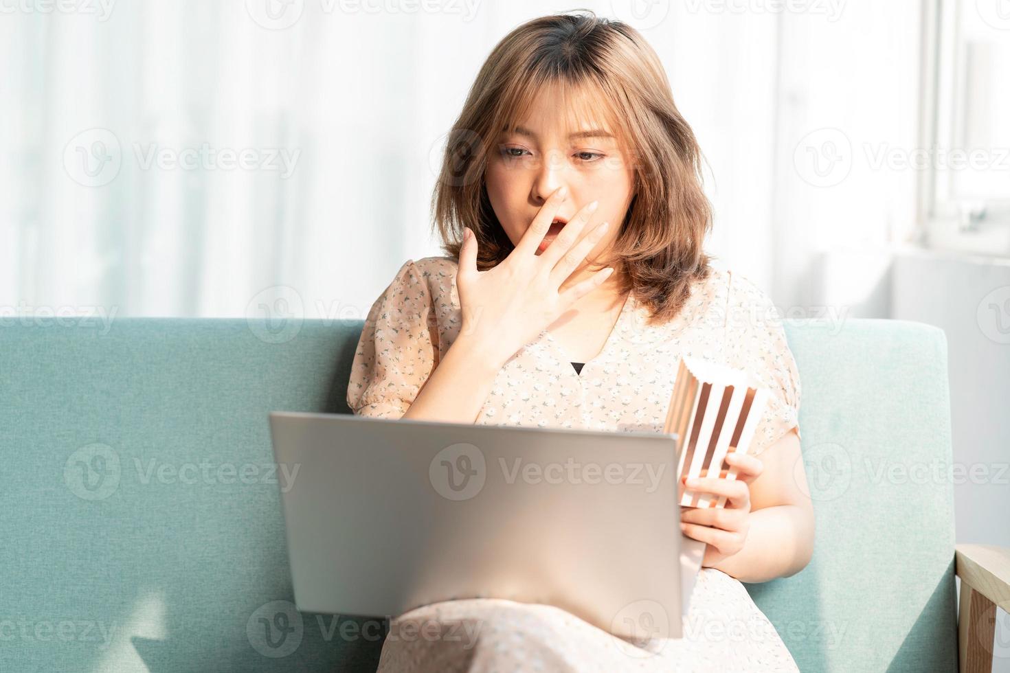 jovem asiática sentada no sofá, comendo e assistindo filmes no laptop foto