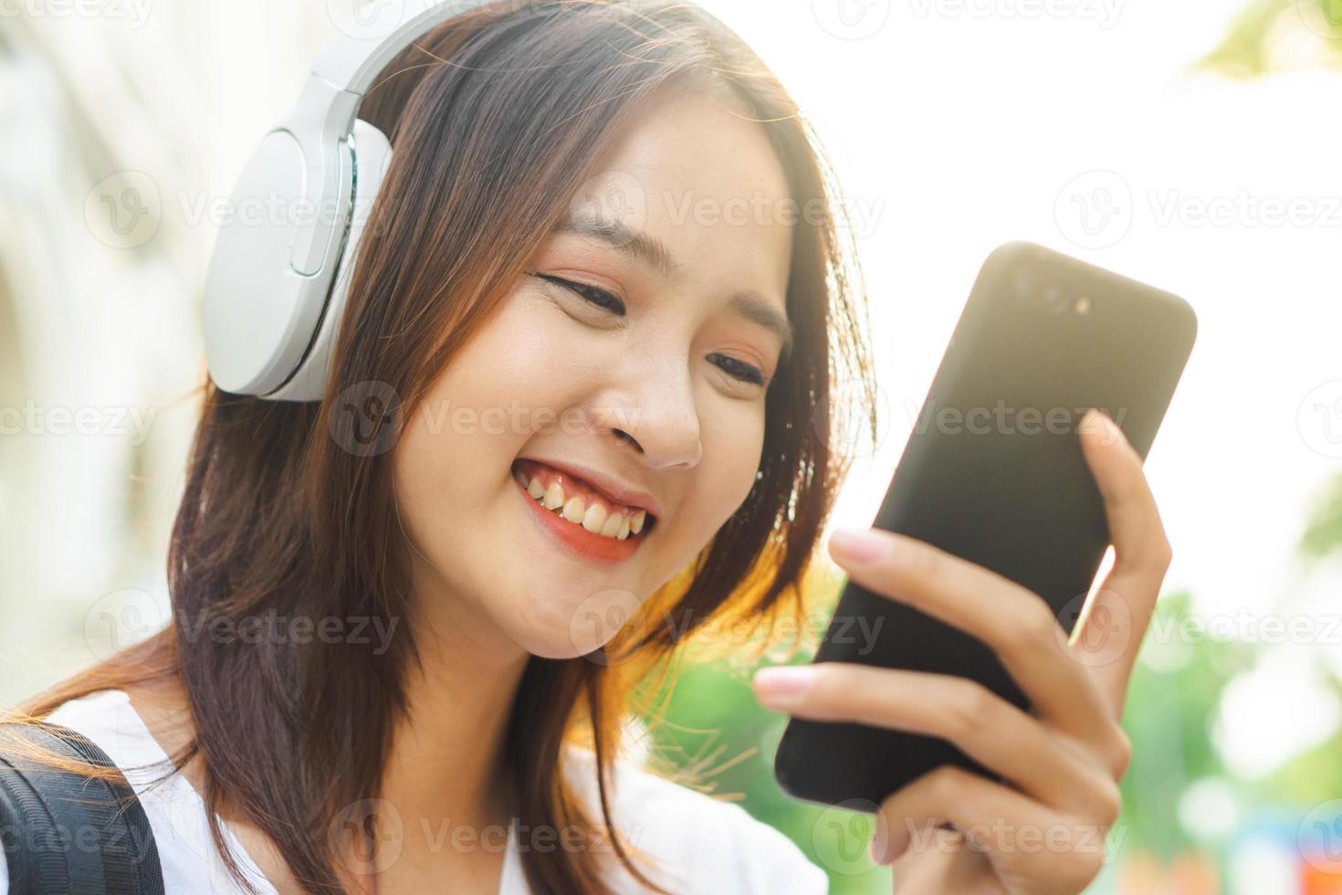 jovem asiática lendo texto em seu telefone e usando fones de ouvido para ouvir música na rua foto