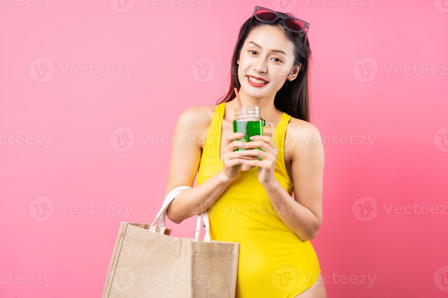 bela jovem asiática em maiô amarelo, bolsa e chapéu de aba larga posando em fundo rosa foto