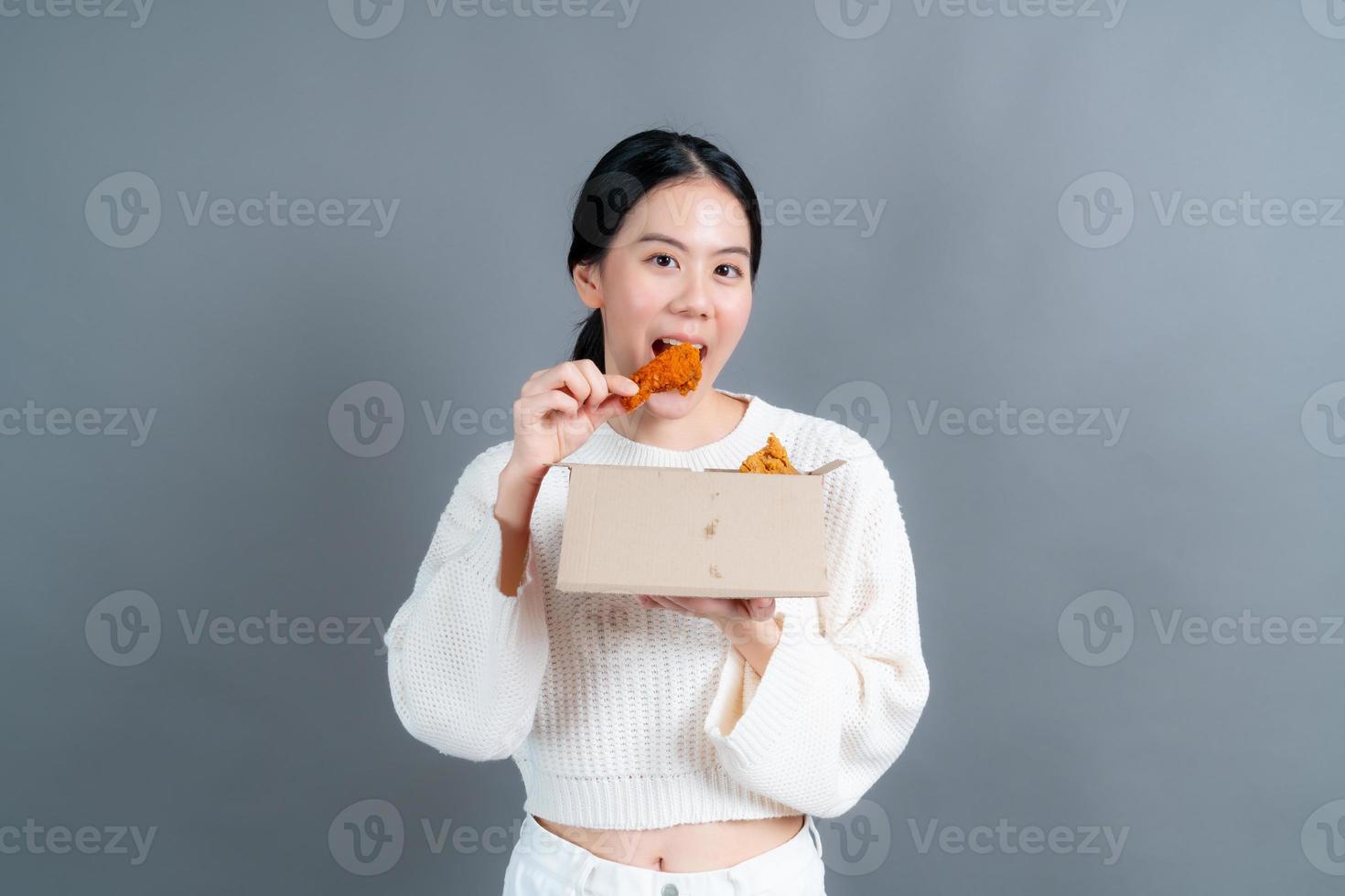 jovem mulher asiática vestindo um suéter com uma cara feliz e gosta de comer frango frito no fundo cinza foto