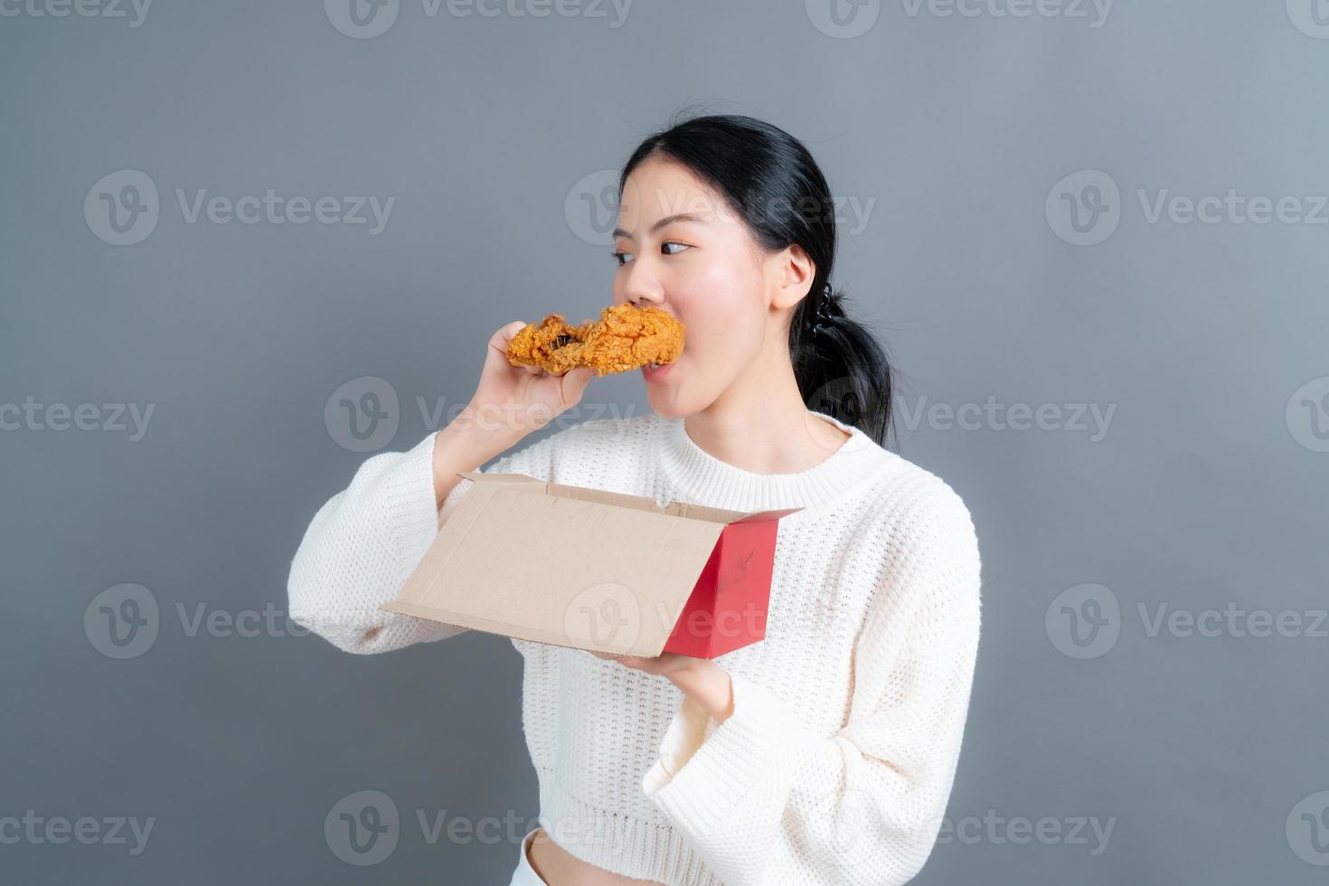 jovem mulher asiática vestindo um suéter com uma cara feliz e gosta de comer frango frito no fundo cinza foto