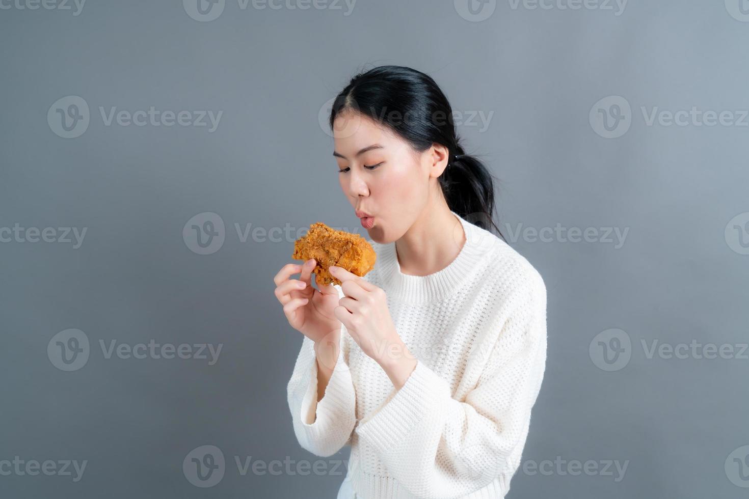 jovem mulher asiática vestindo um suéter com uma cara feliz e gosta de comer frango frito no fundo cinza foto