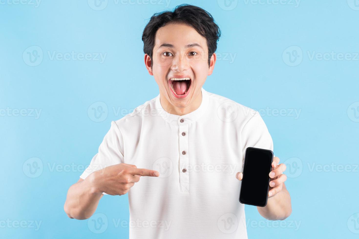 jovem sorridente segurando um smartphone moderno na mão e apontando o dedo na tela branca em branco, sobre fundo azul. foto