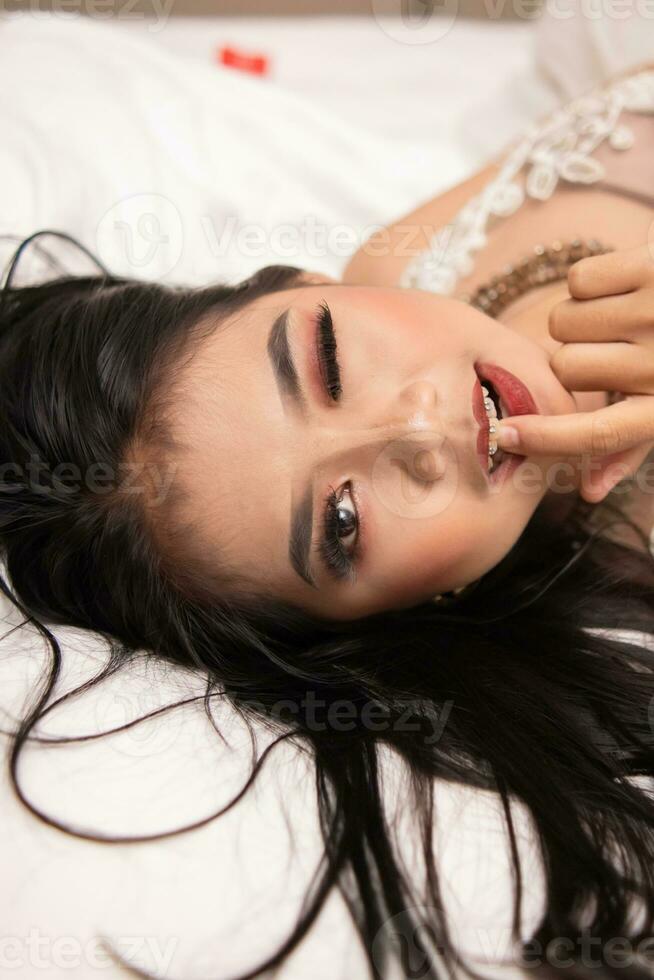 a ásia mulher com Preto cabelo morder dela dedos com uma com tesão expressão enquanto dormindo em uma branco cama foto