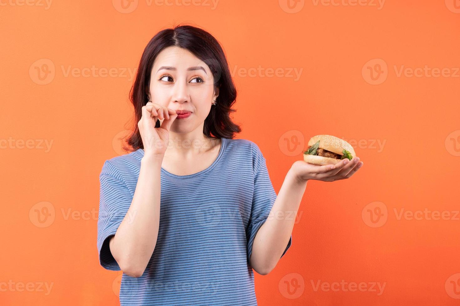 jovem mulher asiática comendo hambúrguer em fundo laranja foto