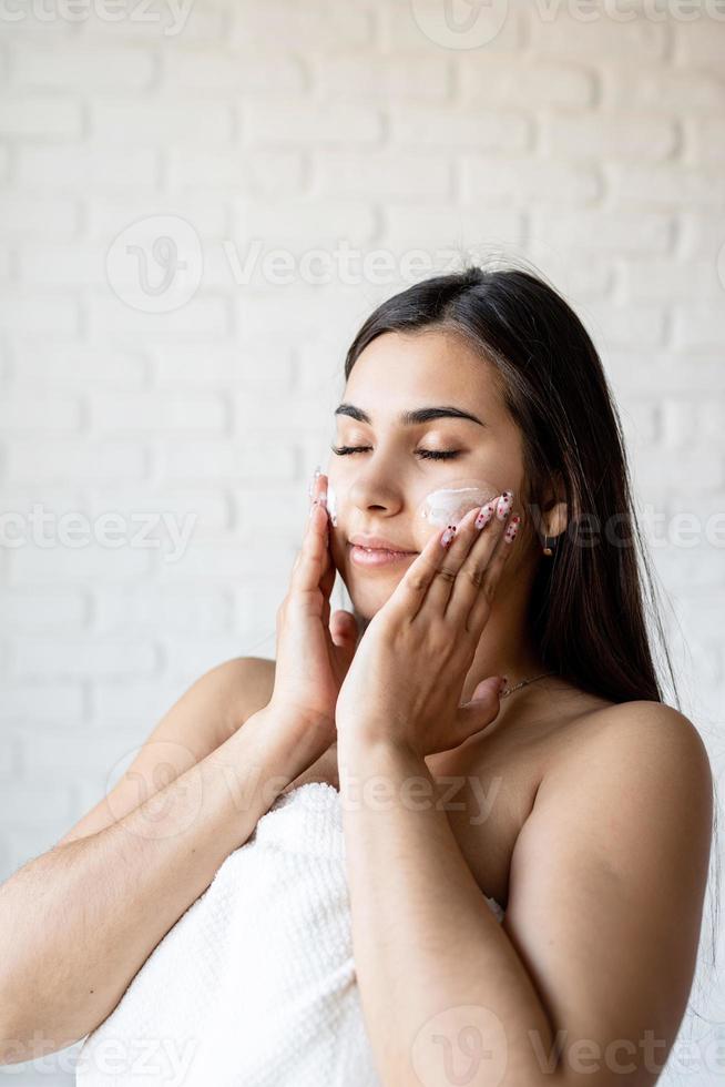 Mulher bonita feliz vestindo roupão de banho e aplicando creme facial foto