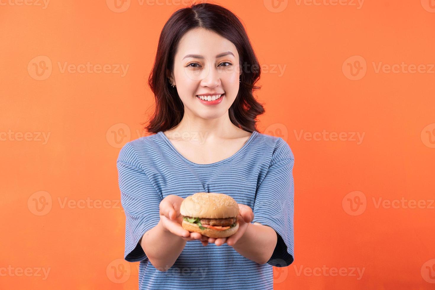 jovem mulher asiática segurando hambúrguer em fundo laranja foto