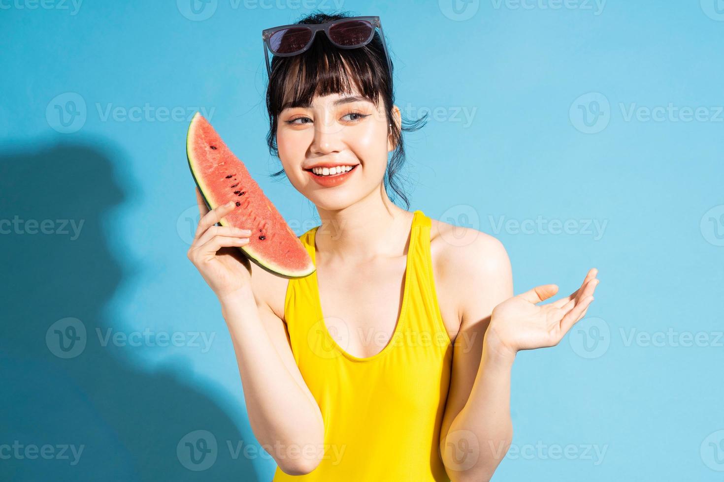 linda mulher asiática vestindo macacão amarelo sobre fundo azul e comendo frutas tropicais, conceito de verão foto