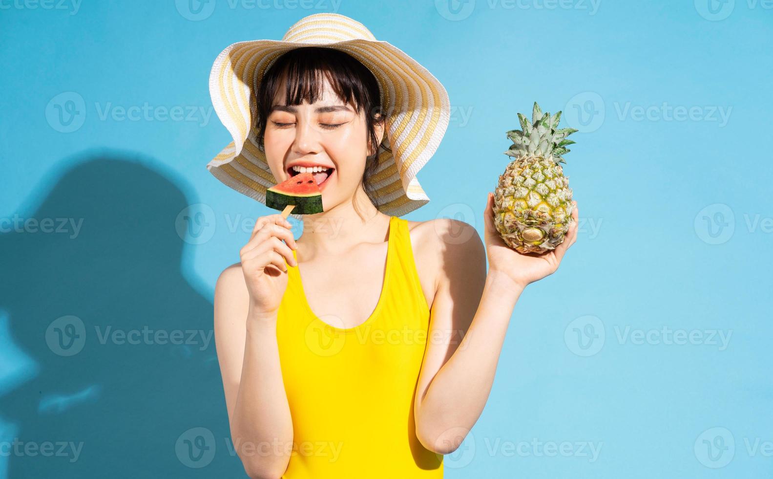 linda mulher asiática vestindo macacão amarelo sobre fundo azul e comendo frutas tropicais, conceito de verão foto