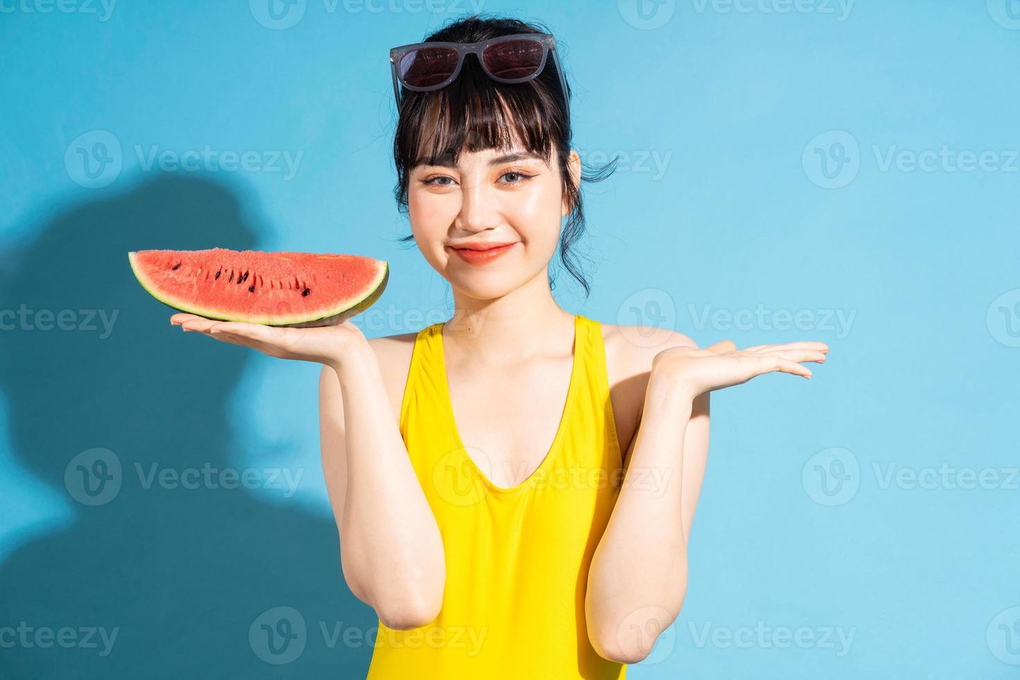 linda mulher asiática vestindo macacão amarelo sobre fundo azul e comendo frutas tropicais, conceito de verão foto