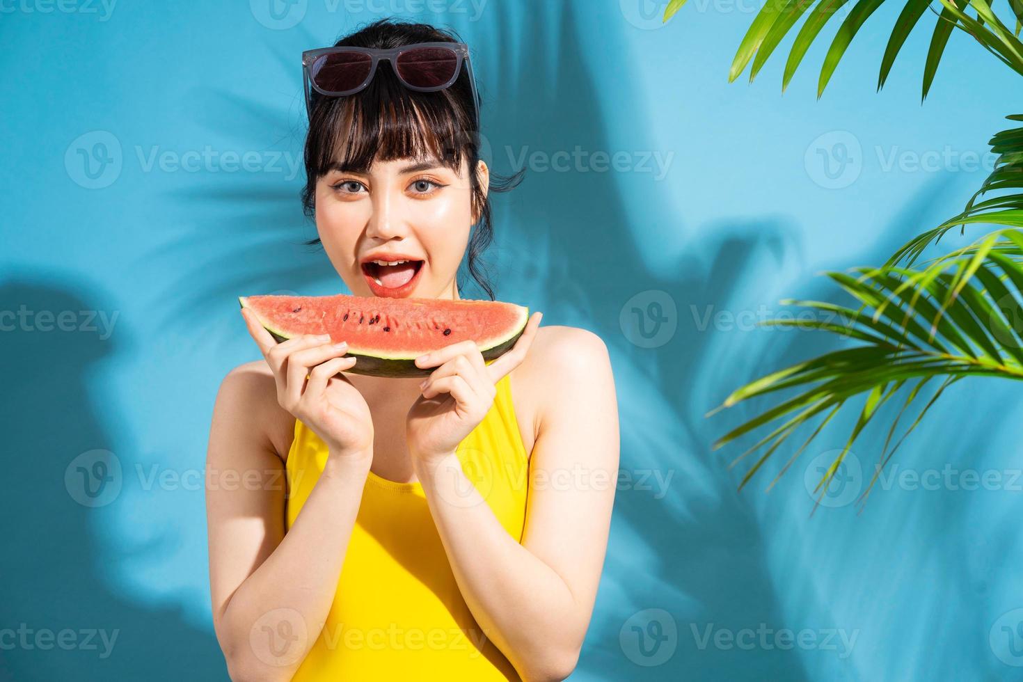 linda mulher asiática vestindo macacão amarelo sobre fundo azul e comendo frutas tropicais, conceito de verão foto