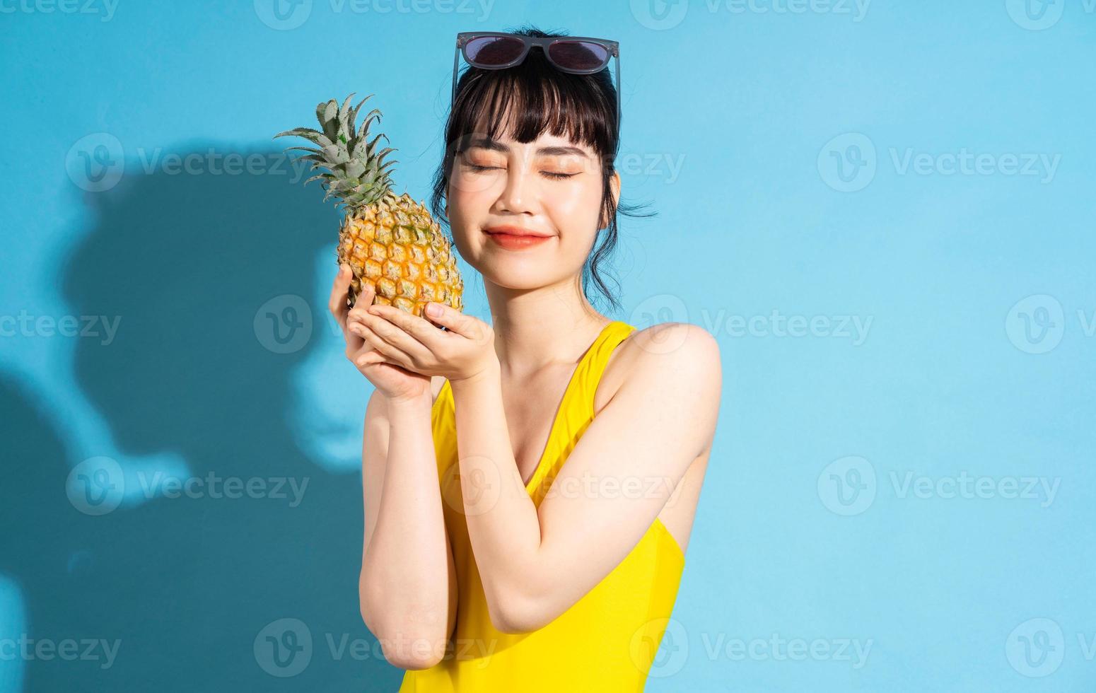 linda mulher asiática vestindo macacão amarelo sobre fundo azul e comendo frutas tropicais, conceito de verão foto